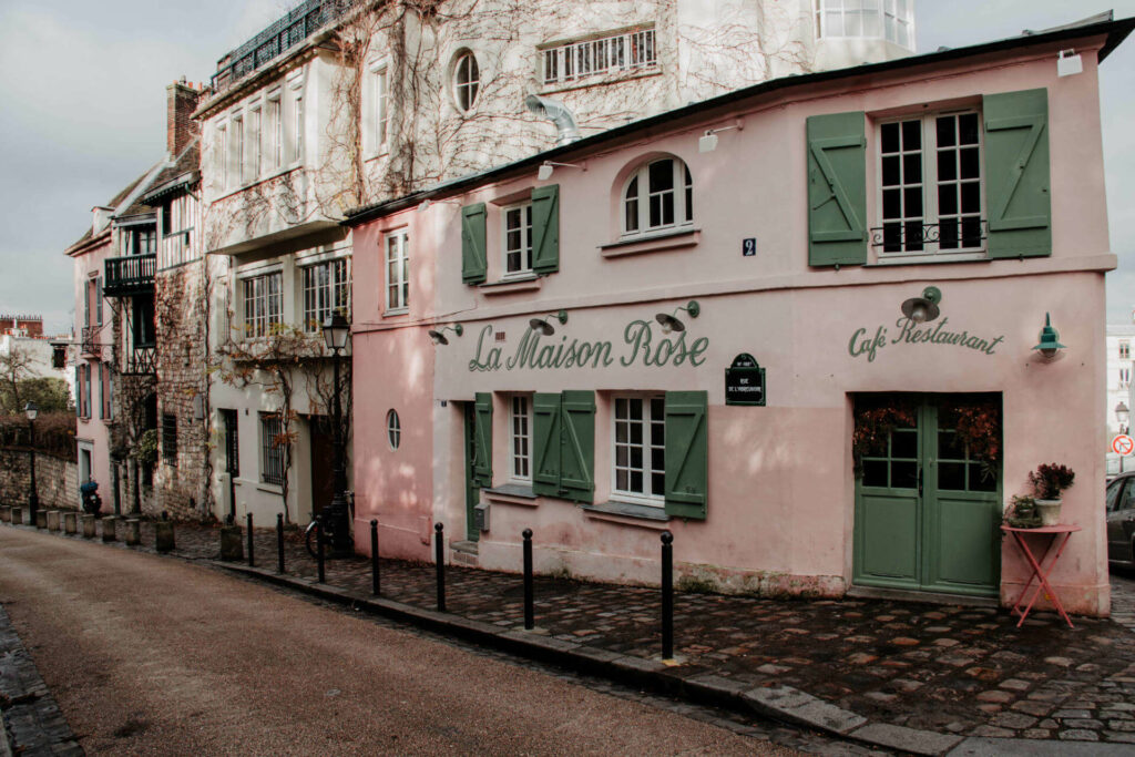 La Maison Rose ist ein Pariser Café und Restaurant in einer kleinen Straße mit rosa Fassade und grünen Fensterläden.