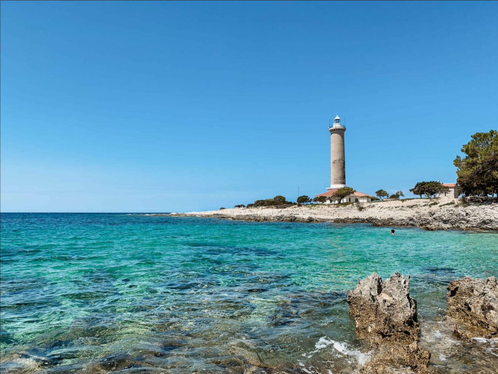 Türkisblaues Meer auf der Insel Dugi Otok vor der Küste Kroatien lädt zum Schnorcheln ein, in der Ferne ragt ein Leuchtturm in den Himmel.