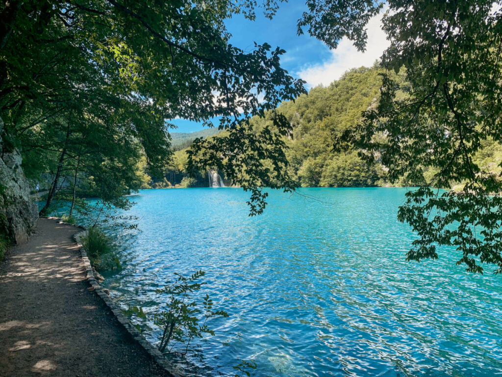Neben einem glasklaren, türkisfarbenen See im Nationalpark Plitvicer Seen in Kroatien führt ein unter Bäumen liegender Wanderweg entlang.