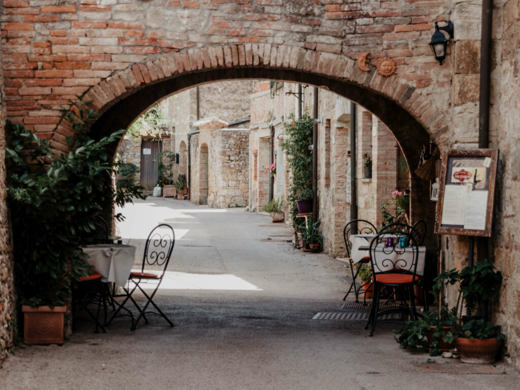 Im Schatten des Torbogens einer Mauer im italienischen Ort San Quirico d’Orcia stehen Tisch und Stühle des Retaurants.