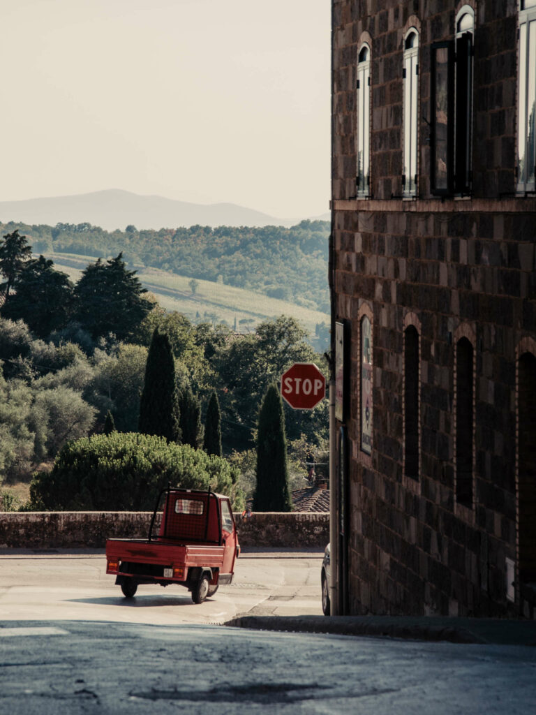 Eine rote dreirädrige Vespa, in Italien als Ape bekannt, biegt hinter einem Gebäude in eine Straße ein. Im Hintegrund leuchtet das satte Grün der toskanischen Zypressen, die jeden Toskana-Urlauber verzaubern.