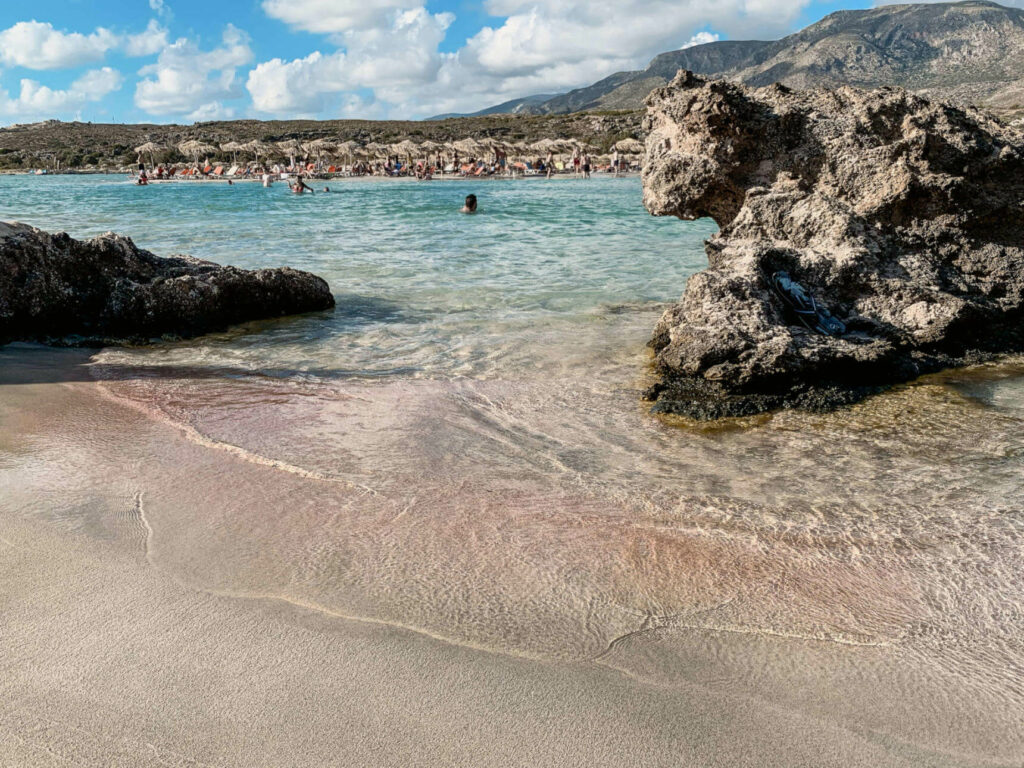 Sandstrand in Elafonissi auf Kreta gesäumt von Felsen, im Hintergrund liegen Besucher unter Strohsonnenschirmen oder schwimmen im Meer.