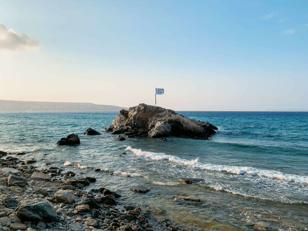 Leichter Wellengang an einem Steinstrand, aus dem türkisblauen Wasser erhebt sich ein Felsen mit Griechenlands Flagge darauf.