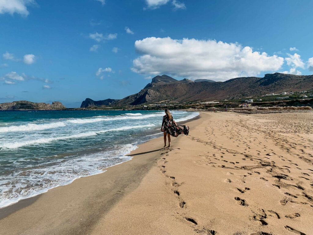 Fußabdrücke einer Frau an dem langen, goldfarbenen Sandstrand Falassarna auf der griechischen Insel Kreta. Die Frau spaziert mit wehendem Kleid an den hereinbrechenden Wellen vorbei.