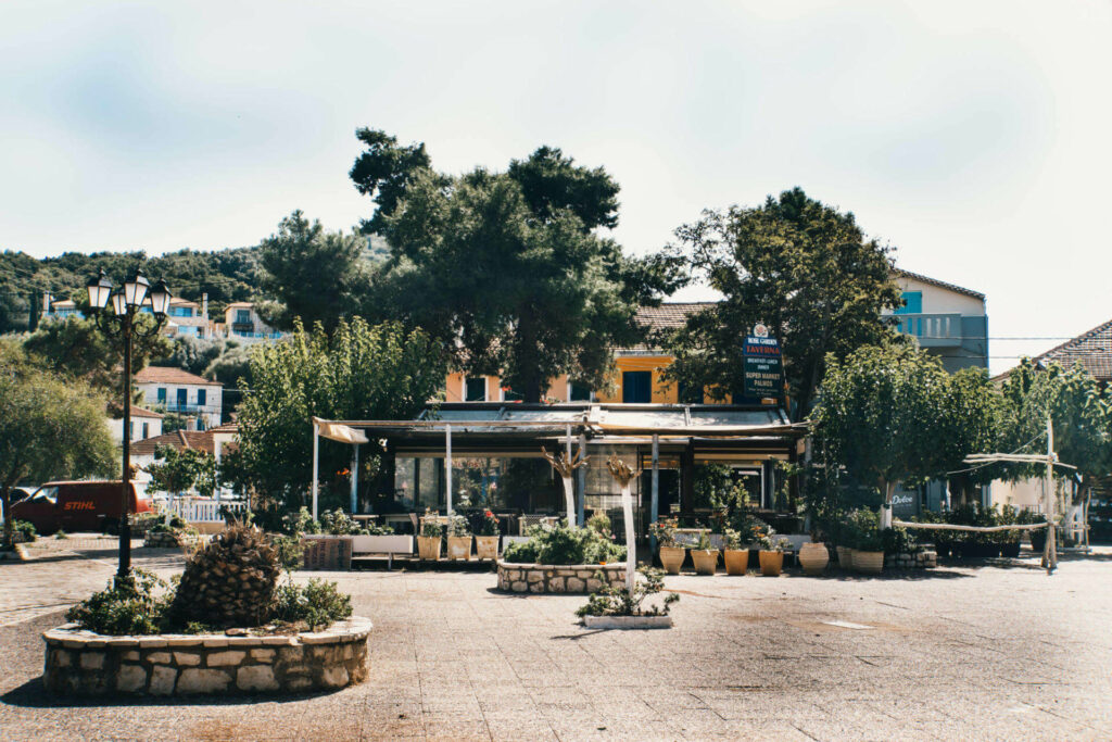Ein gepflasterter Platz auf der Insel Meganisi, ein gelbes Haus ist hinter vielen grünen Bäumen zu erkennen.