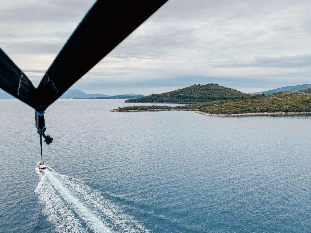 Ein Parasailing-Boot hinterlässt Fahrspuren im Meer, über ihm bauen sich die Flügel des Parasailing-Gefährtes auf, im Hintergrund die Weite des Meers und die Ausläufer von Lefkada, der kleinen griechischen Insel.