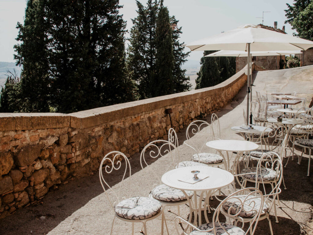 Die Sonnen-Terrasse einer Bar lädt mit weißen Stühlen in verschnörkeltem Design und Sonnenschirme zu einer Aussicht auf die bezaubernde toskanische Landschaft mit ihren Zypressenbäumen ein.