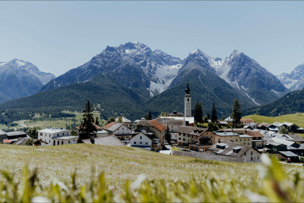 Blick über eine Wiese hinein in ein Dorf mit Kirchturmspitze, dahinter Bergpanorama, in Ftan in der Schweiz.