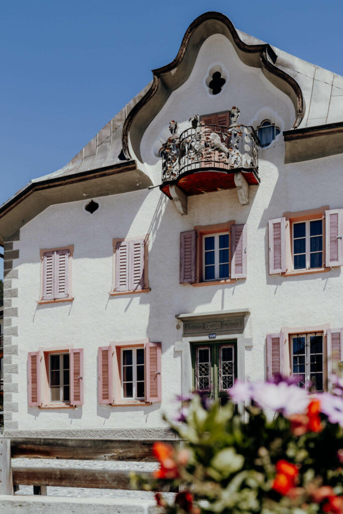 Ein weißes Haus im Jugendstil mit rosa Fensterläden und einem verschnörkelten Balkon im zweiten Stock, in Sent, Schweiz.
