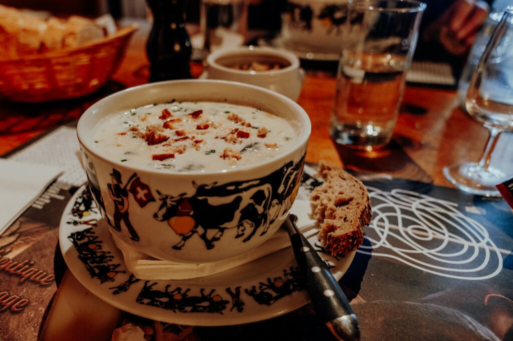 Eine Porzellanschüssel mit Kuhmotiv und geschmolzenem Käse und Croutons darin steht auf einem Teller angerichtet, auf dem ein Stück einer Scheibe Brot liegt.