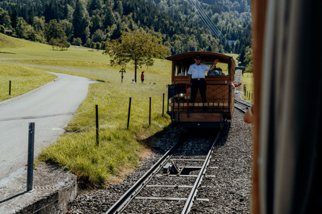 Eine Oldtimer-Standseilbahn fährt über Gleise auf Kies, im Hintergrund liegt ein Wald, links neben der Bahn verläuft ein Weg.