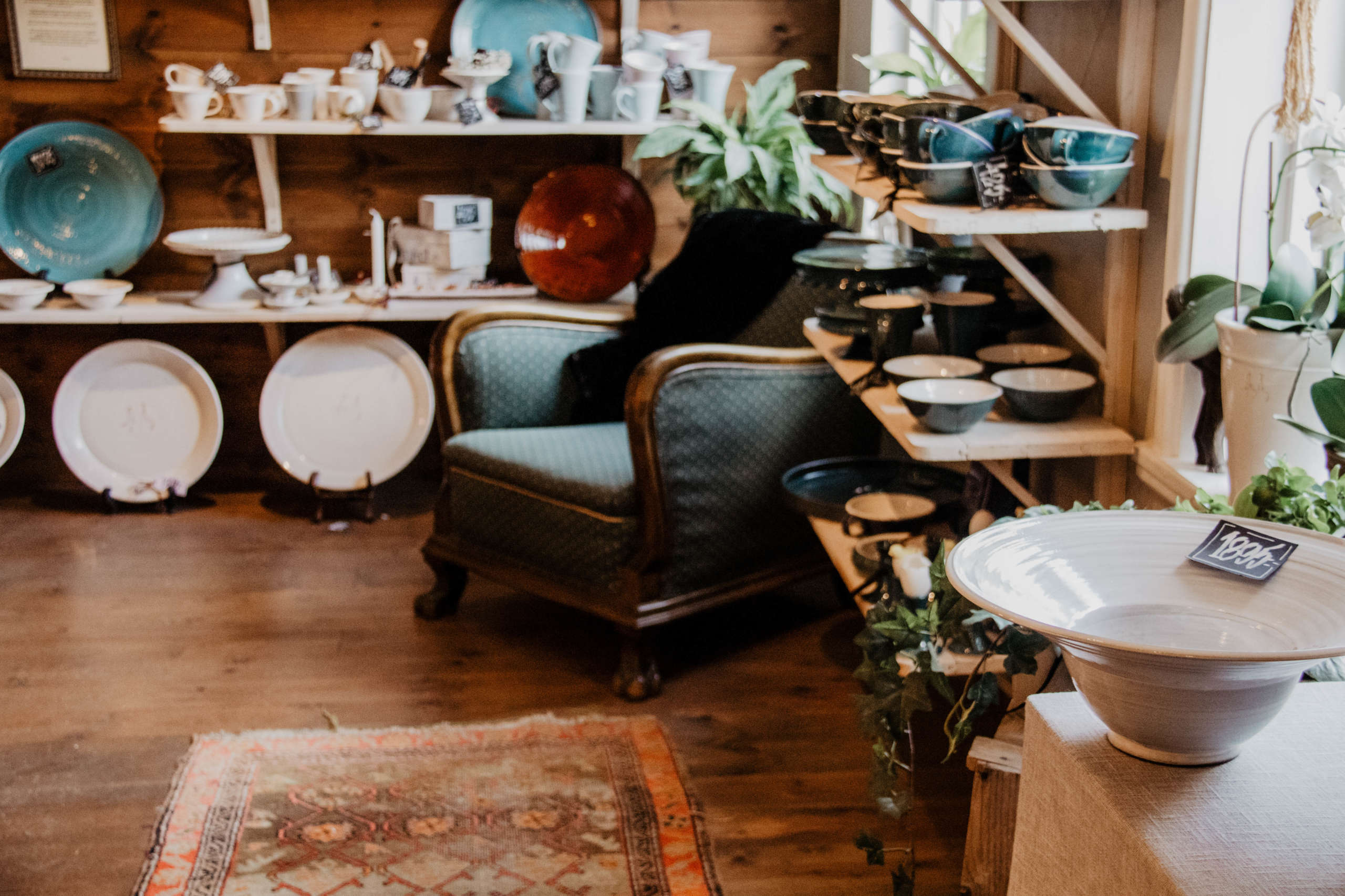 In einem kleinen Geschäft mit Holzboden steht ein großer dunkelbrauner Holzsessel mit grünem Polster, um ihn herum türmen sich allerlei Keramikwaren wie Teller, Tassen und Schüsseln auf.