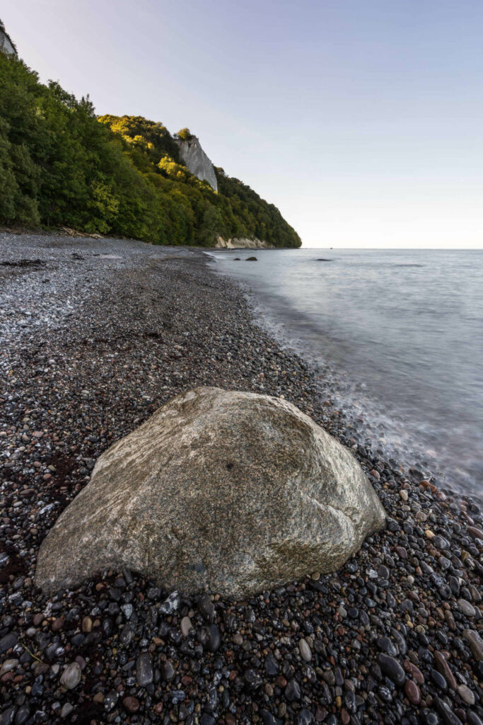 Der Königsstuhl mit seinem weißen Kreidefelsen liegt in der Ferne auf einem bewaldeten Felsen, das Meer schlägt an den Kiesstrand, im Vordergrund befindet sich ein großer Stein.
