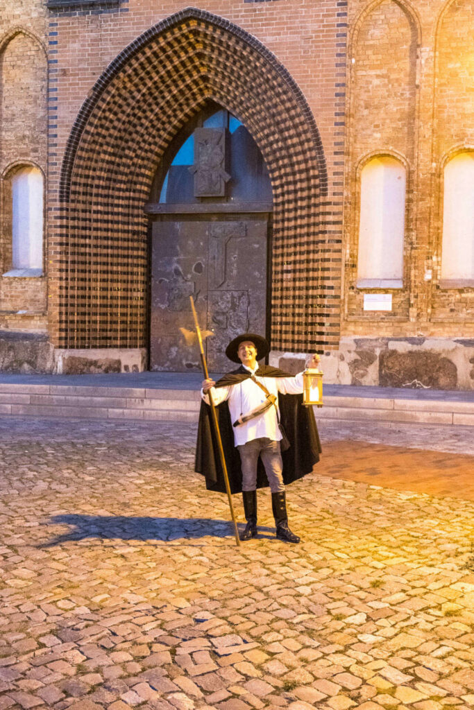 Ein Nachtwächter steht im mittelalterlichen Kostüm und mit einer Öllampe in der Hand vor einer großen Kirchentür in Rostock. Er zeigt den Besuchern die Sehenswürdigkeiten der Stadt.