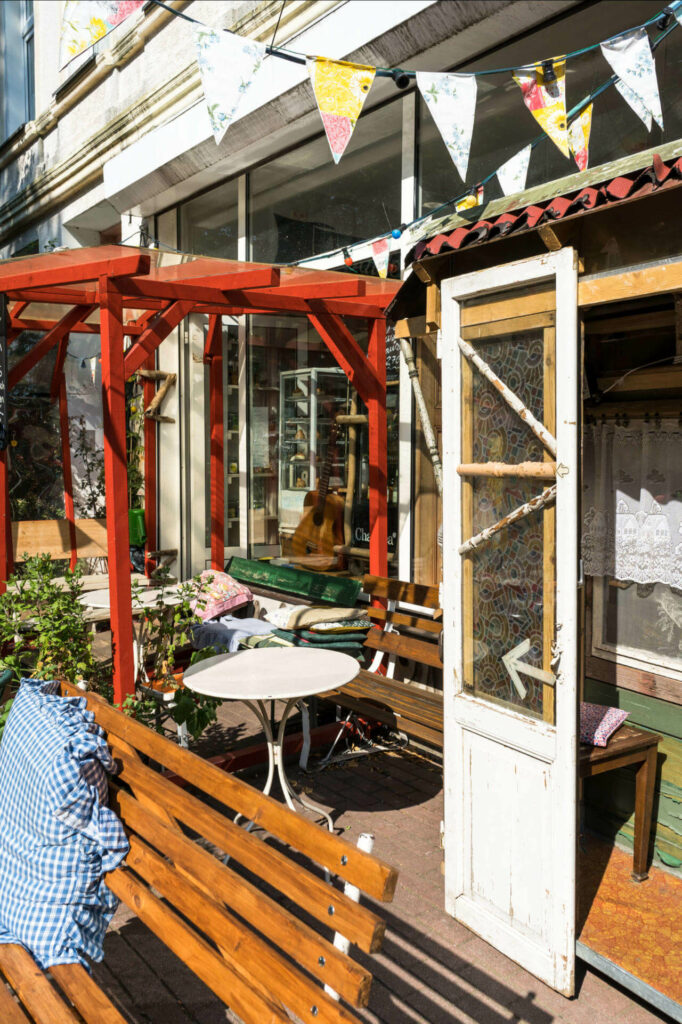 Den Eingang eines kleinen, alternativen Cafés in Rostock in der Kröpeliner-Tor-Vorstadt ziert eine rote Pergola, Holzbänke, Kissen und weiße Tische, eine alte weiße Holztür mit Pfeil zeigt ins Café hinein.