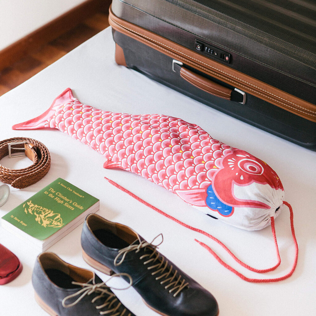 Der praktische Wäschesack für Reisen in Form eines roten Koi-Karpfens liegt neben dem Reisekoffer, Schuhen und einem Buch.