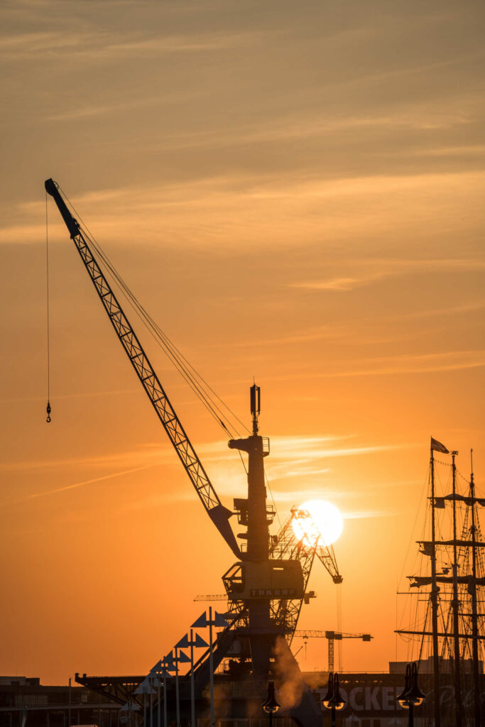 Ein alter Kran im Rostocker Hafen im Licht der untergehenden Sonne, der Himmel ist komplett orange gefärbt und wirft sein Licht auf den Kran.
