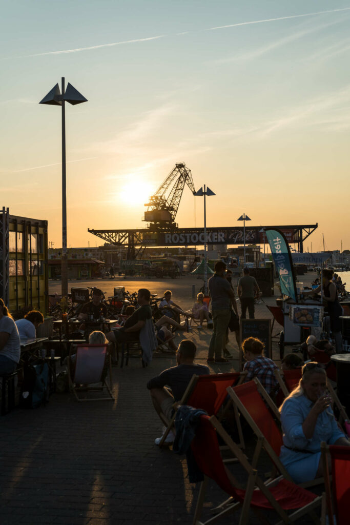 Im Sommer einen Besuch wert: Menschen sitzen in einer Bar am Hafen von Rostock bei Sonnenuntergang, im Hintergrund befindet sich ein Schiffskran.