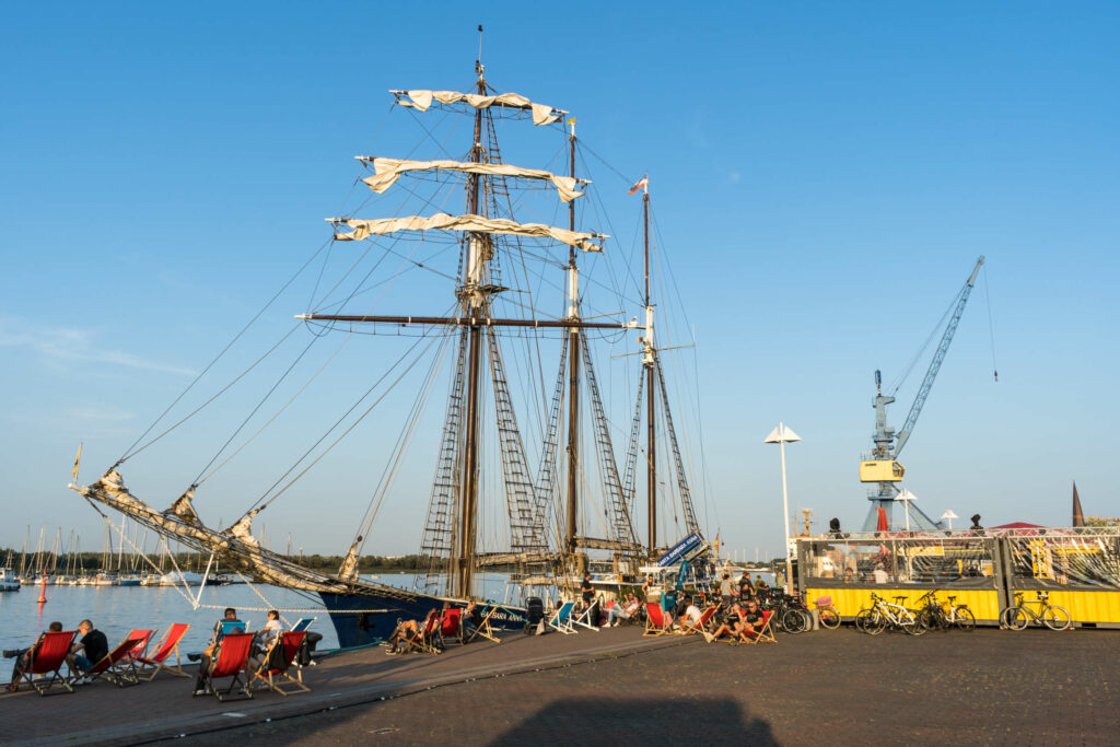 Ein umfunktioniertes Schiff liegt in Rostock an einem Dock an, davor sitzen Menschen in Strandstühlen, daneben befindet sich ein kleiner Unterstand mit anlehnenden Fahrrädern, der Himmel ist strahlend blau, man blickt auf einen Kran.