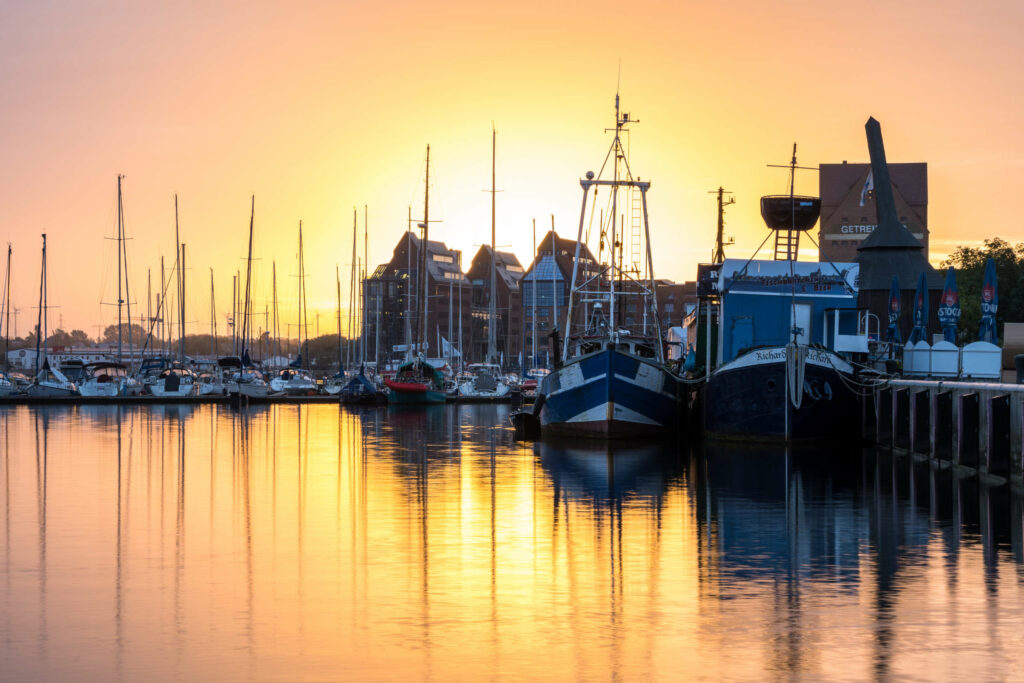 Diverse Schiffe, Yachten und Dampfer liegen bei Sonnenaufgang im Rostocker Stadthafen. die untergehende Sonne spiegelt sich im Wasser.