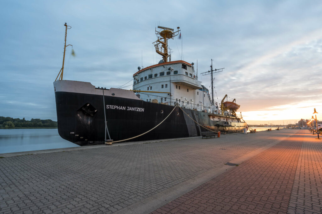 Die Stephan Jantzen, ein riesiger Eisbrecher, liegt im Hafen von Rostock, im Hintergrund geht die Sonne auf, das Schiff dockt an eine gepflasterte Plattform, die mit Straßenlaternen gesäumt ist, an.