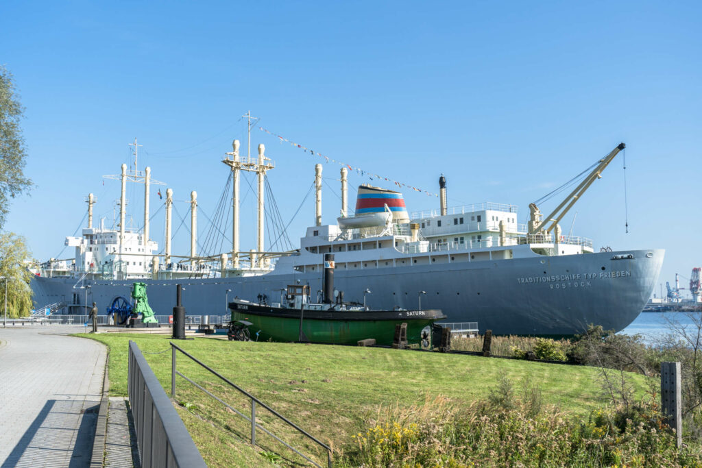 Das Traditionsschiff Typ Frieden liegt am Ufer von Rostock an, vor ihr liegt ein kleines grünes Boot, davor Gras und gepflasterter Weg, strahlend blauer Himmel.