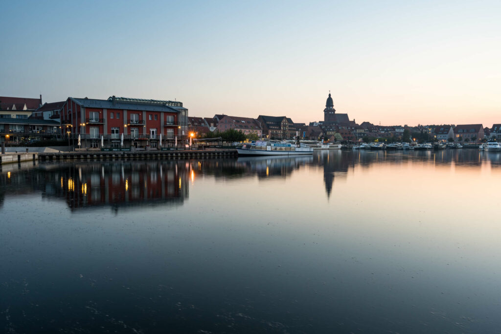 Die Stadt Waren an der Müritz liegt im Sonnenaufgang am Wasser, rote Häuser, eine Kirche und anlegende Schiffe sind in der Dämmerung zu erkennen.