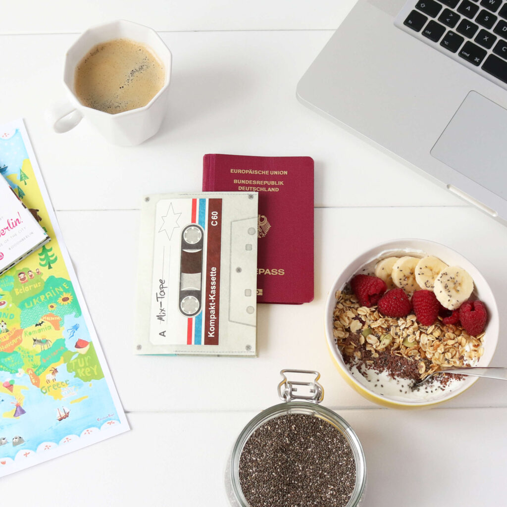 Auf einem Tisch liegen angerichtet eine Schüssel mit Müsli, ein Weckglas mit Chia-Samen, eine Kaffeetasse, ein Laptop, ein Reisepass, sowie die passende Reisepasshülle in Form einer Musikkassette.