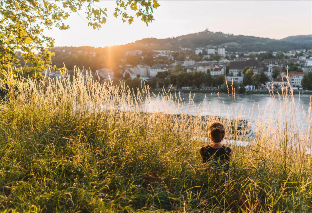 Reisebloggerin Melanie sitzt in hohem Gras und genießt den Sonnenuntergang an der Donau, die durch die sehenswerte Stadt Linz fließt.