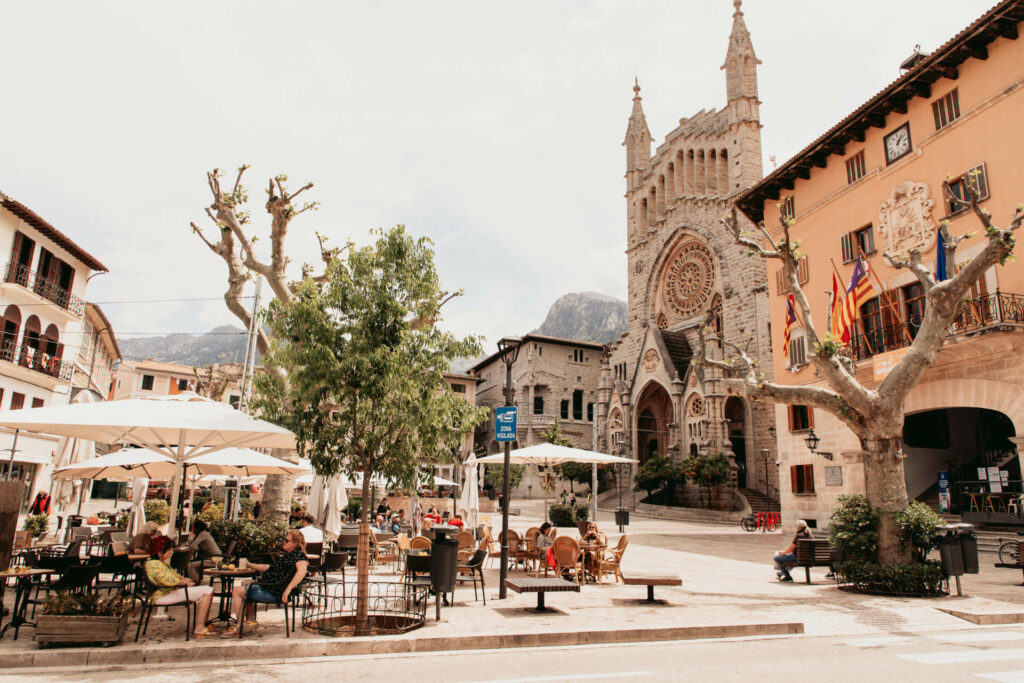 Der belebte Plaça Constitució in Sóller mit Menschen unter Sonnenschirmen und Blick auf die Kirche Sant Bartomeu auf Mallorca.
