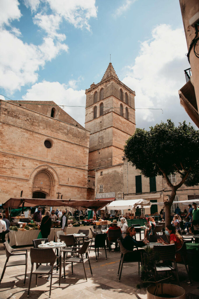 Ein gut besuchtes Café auf dem Wochenmarkt von Sineu auf Mallorca mit einem Gemüsestand und altertümlichen Gebäuden im Hintergrund.