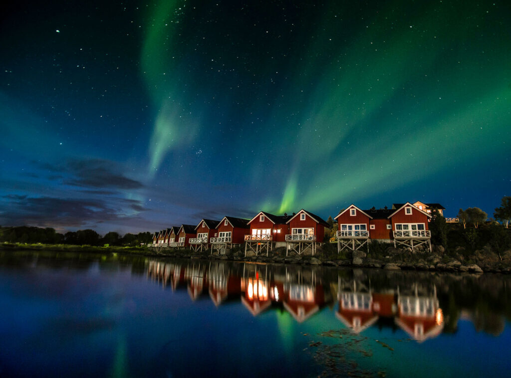 The Northern Lights shine over a row of red shacks by the water at night