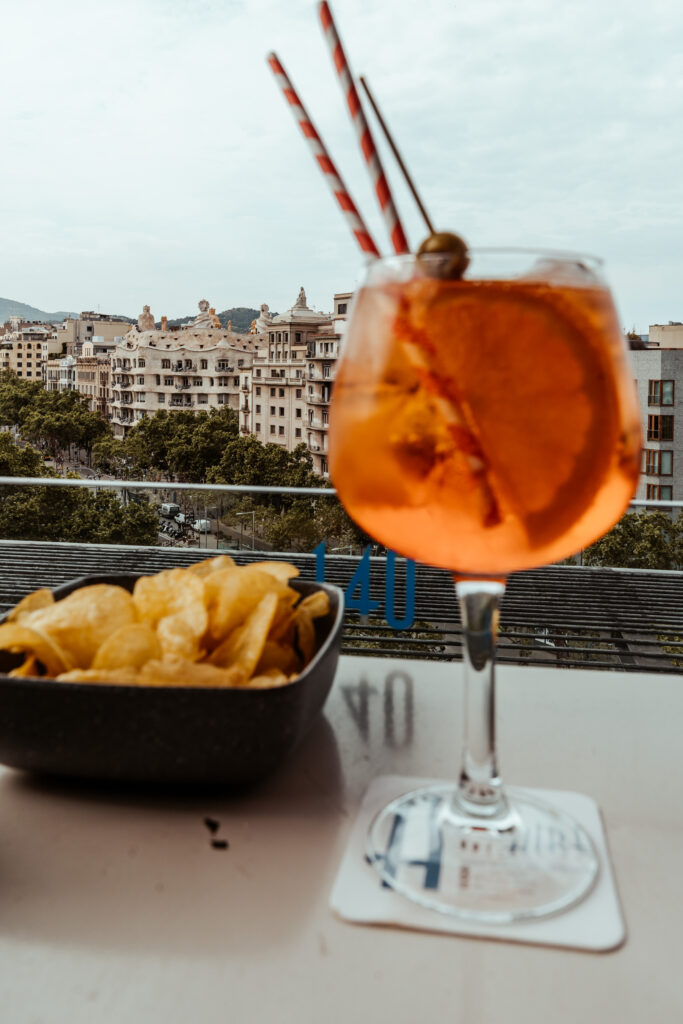 Glas Aperol Spritz und eine Schale Chips stehen auf eine Tisch und man blickt über die Häuser von Barcelona