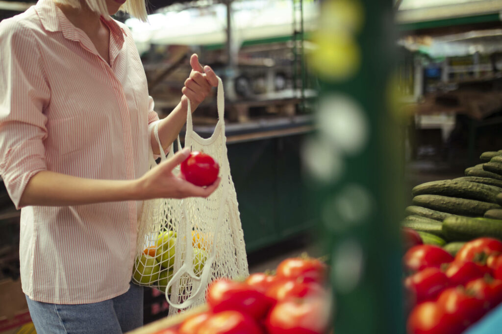 Auf einem Wochenmarkt kauft eine Frau Obst und Gemüse ein und verstaut sie in einem wiederverwendbaren Beutel: Auch auf Reisen ein idealer Begleiter, um nachhaltig unterwegs zu sein.