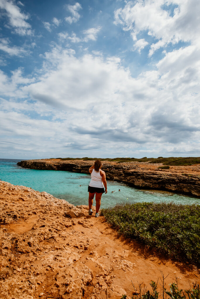 Eine Frau im sommerlichen Outfit steht auf rötlichem Felsgestein in der Bucht Caló d’en Rafalino in S’Illot.