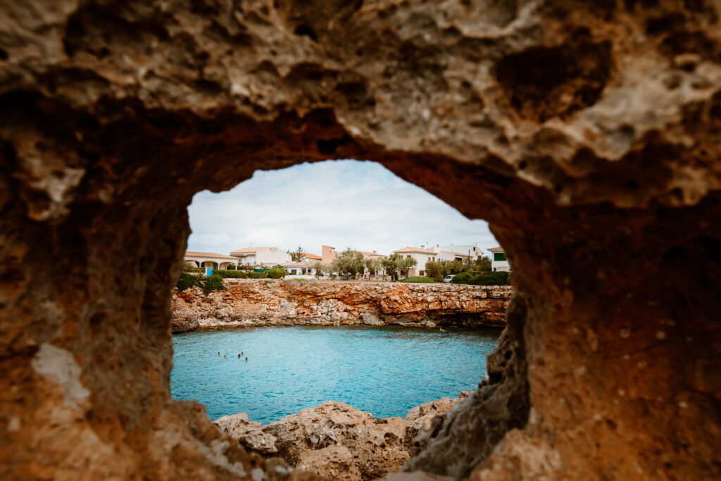 Blick durch ein Lochfenster im Fels auf die Bucht Cala Morlanda mit blauem Meer und felsigen Naturgestein.