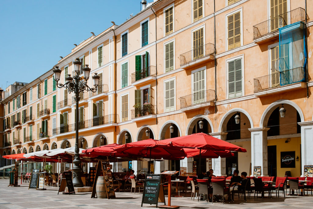 Der große Platz Plaza Mayor mit sandfarbigen Häuserwänden und roten Restaurantschirmen in Palma.