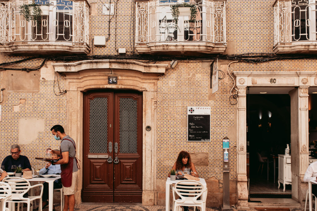 Ein paar Leute sitzen an Tischen vor einer mit Mosaiken besetzen Hauswand der Rua de Santo Antonio in Faro.