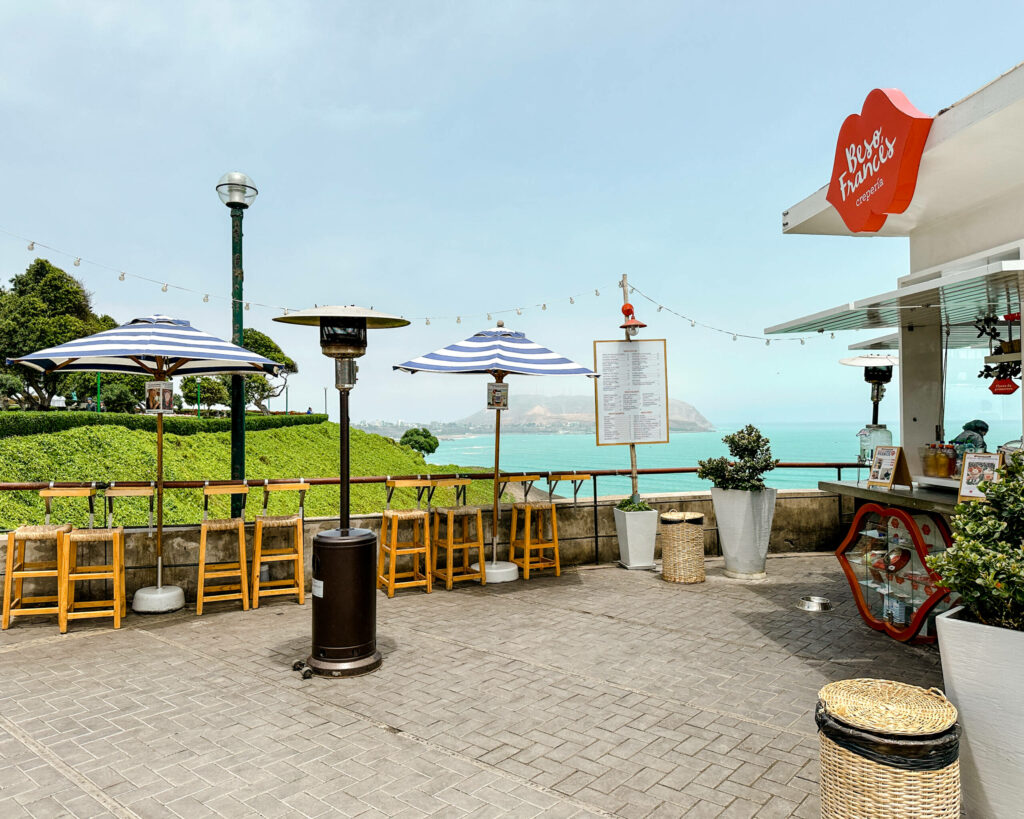 Ein kleines Cafe an der Küstenpromenade von Lima mit blau weißen Schirmen und Blick auf das Meer.