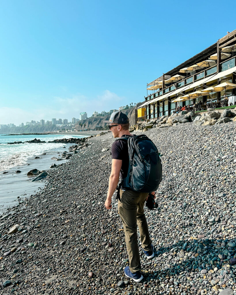 Der kieselsteinige Strand in Lima eignet sich hervorragend zum Spazieren - Reiseblogger Tom von traveloptimizer ist mit Rucksack an der Promenade unterwegs.
