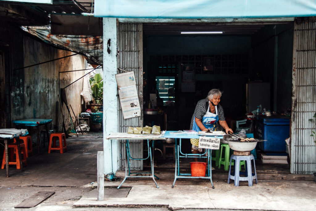 Eine ältere Dame steht hinter einer Streetfood Garküchen in Bangkok und kocht.