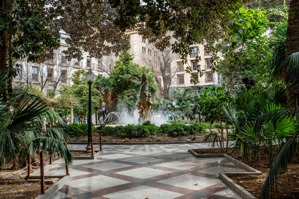 Ein Brunnen inmitten der Altstadt von Alicante zwischen vielen Bäumen und Palmen.