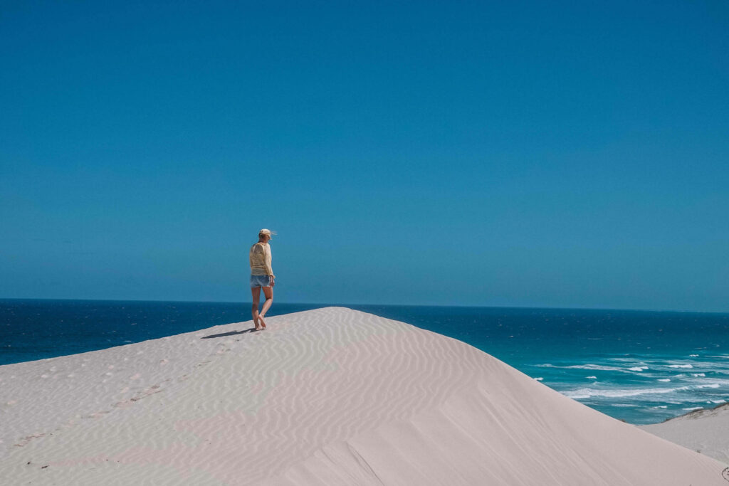 Bloggerin Nina steht auf einer Düne in weißem Sand mit türkisblauem Meer und einem blauen Himmel im Hintergrund.