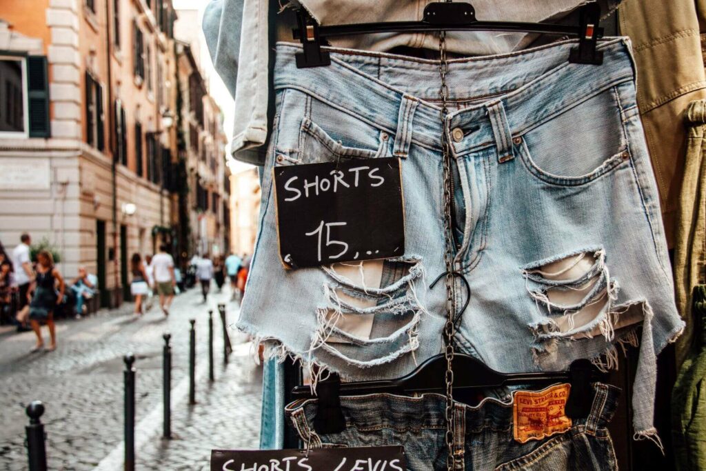 Im Vordergrund hängt eine kurze Jeansshort mit Preisschild, im Hintergrund laufen Menschen durch die Straßen des Viertels Monti in Rom.