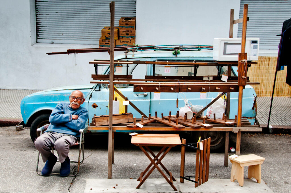 An einem Flohmarktstand auf dem Trödelmarkt Feria Tristan Narvaja in Montevideo sitzt ein fröhlicher Verkäufer vor seinem Flohmarktstand mit Holzerzeugnissen.