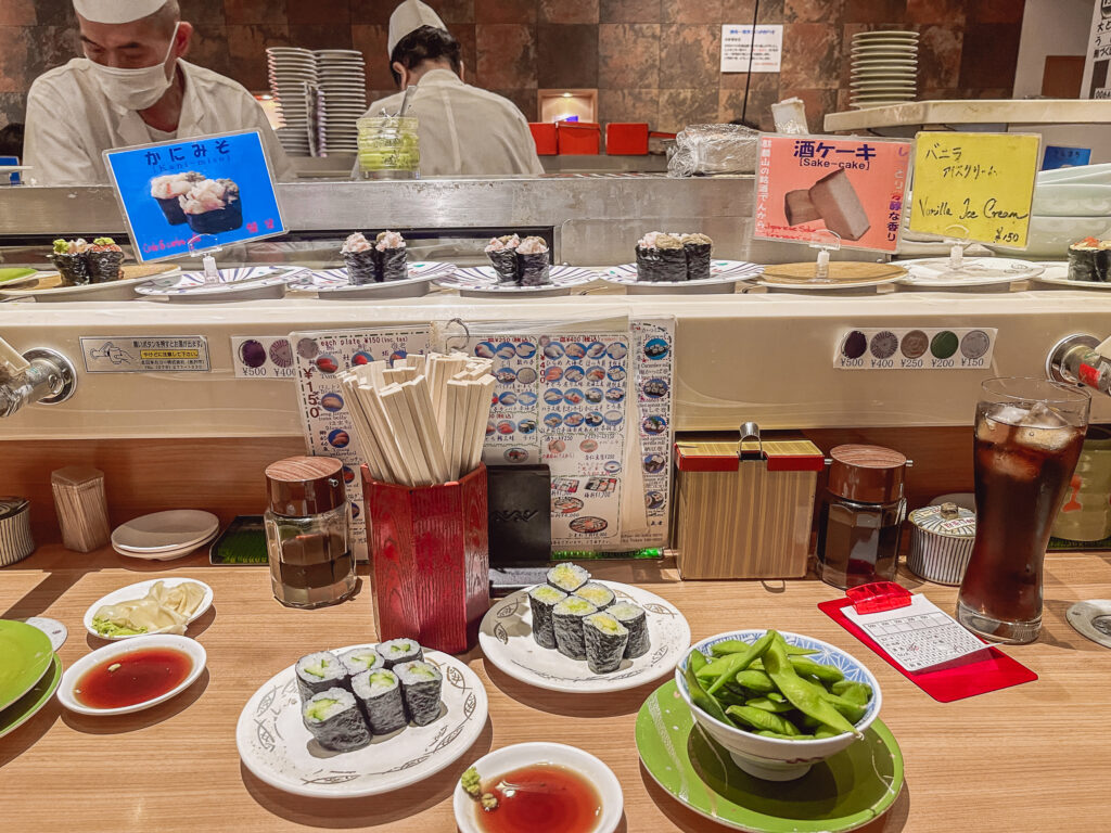 Blick auf ein Running Sushi Band, auf einem Tisch stehen kleine Teller mit Sushi Rollen und Edamame,.