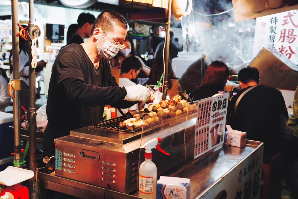 Ein Mann steht hinter einem kleinen Grill auf dem Austernpilze liegen auf dem Shilin Nachtmarkt in Taipeh.